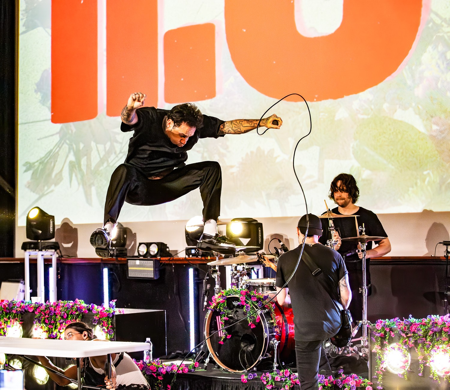 Performer jumps on stage with a microphone, two musicians playing instruments, and a large screen in background.