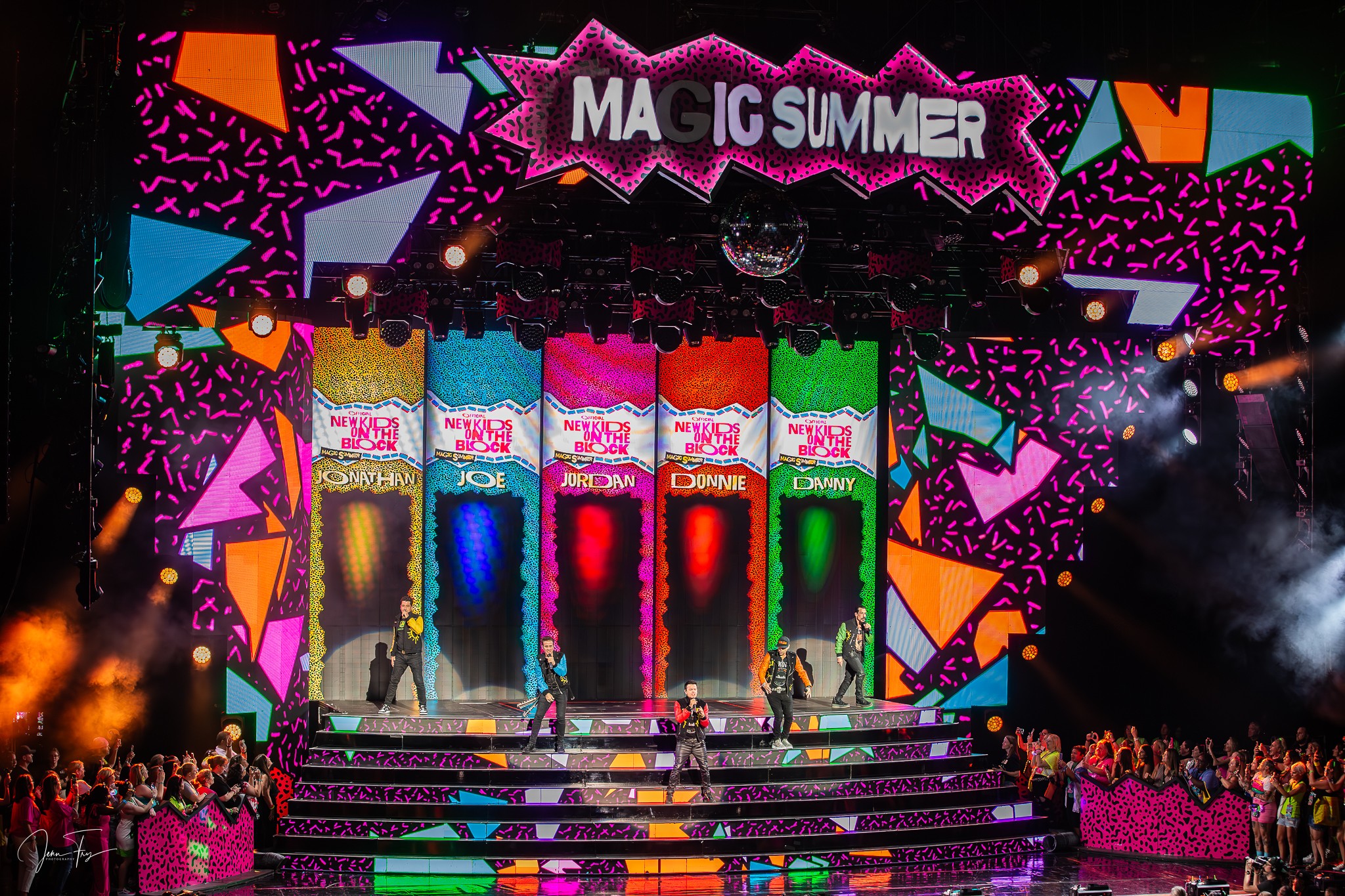 Stage with performers and colorful backdrop reading 'MAGIC SUMMER', surrounded by an audience, bright lights, and vibrant decorations.