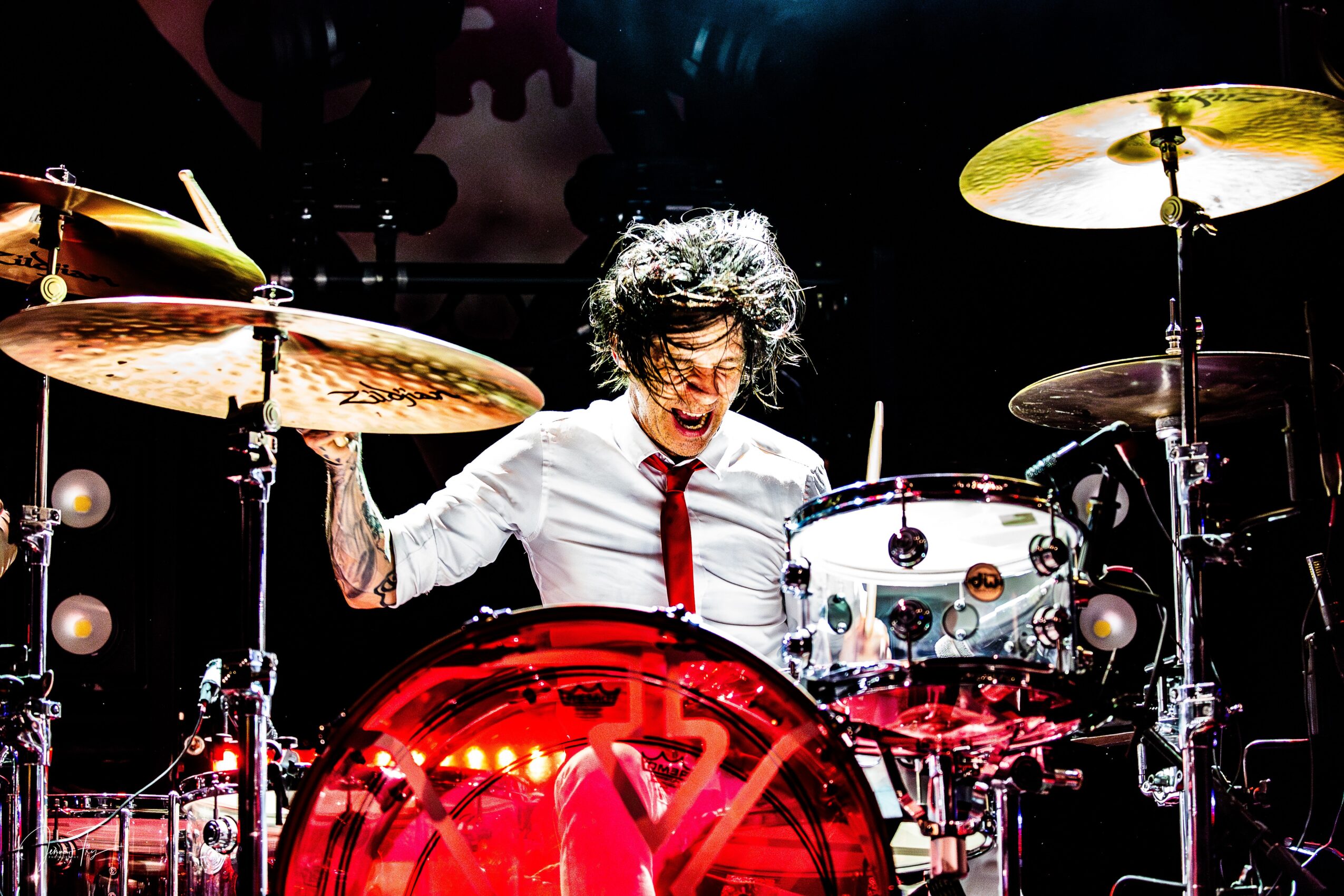 Person playing drums on stage, smiling, with drumsticks, surrounded by cymbals and drums, wearing a white shirt and red tie.