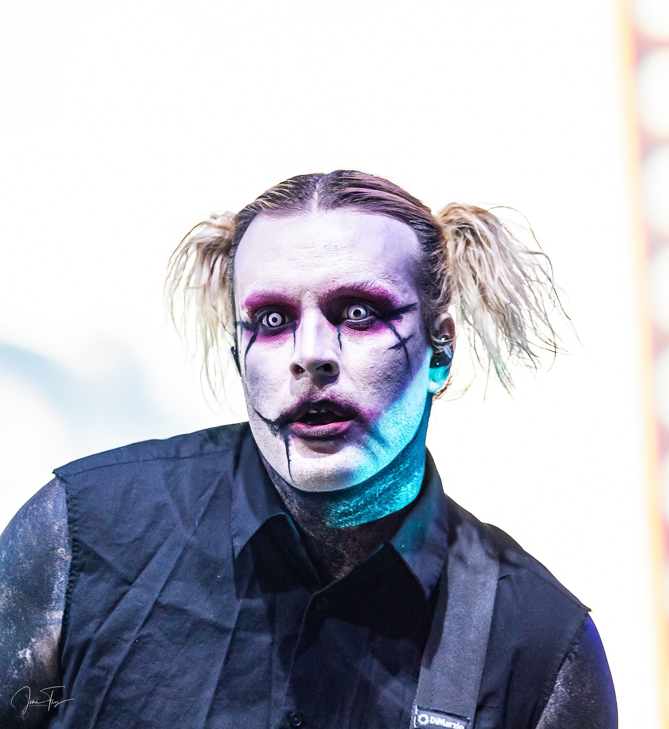 Person with clown makeup, pale face, dark eyes, and messy hair in pigtails, wearing dark clothing, against a bright background.