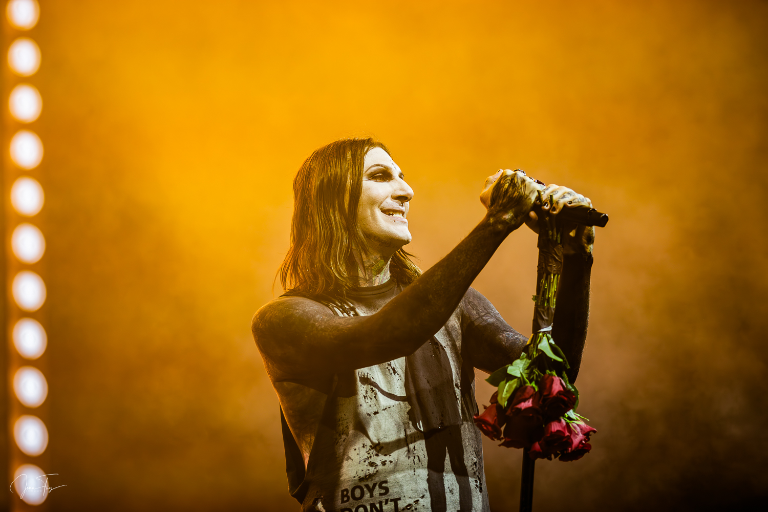 A woman with dark hair holds a bouquet of pink roses on stage with yellow lighting behind her.