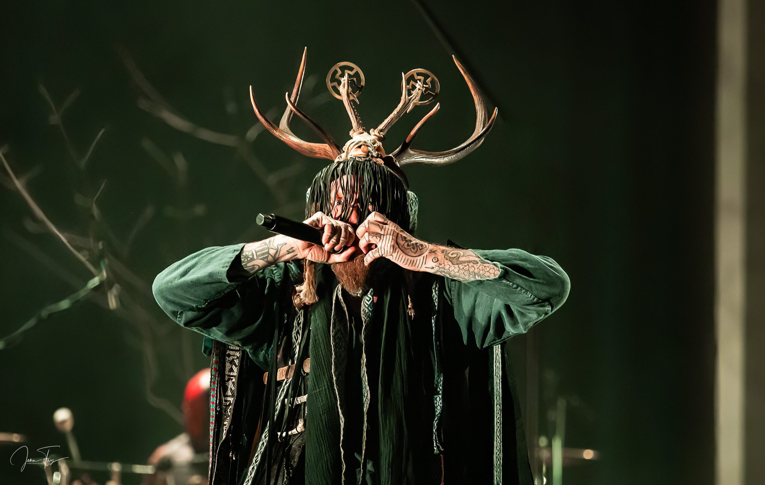 Person wearing a horned mask and green clothing, holding a prop or instrument, on stage with dark background.