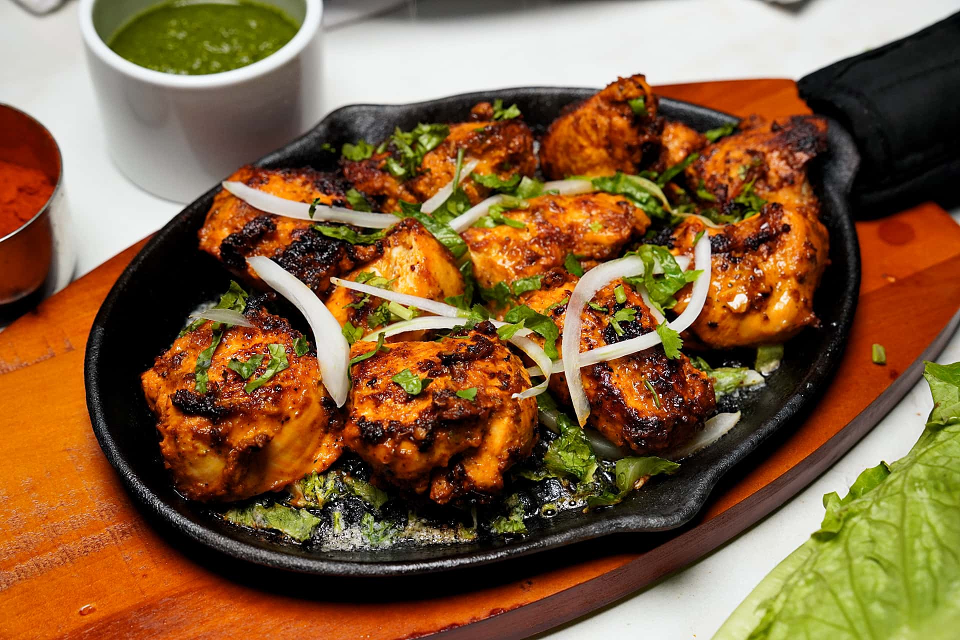 Grilled chicken pieces garnished with sliced onions on a sizzling black plate, with green and red sauces nearby.