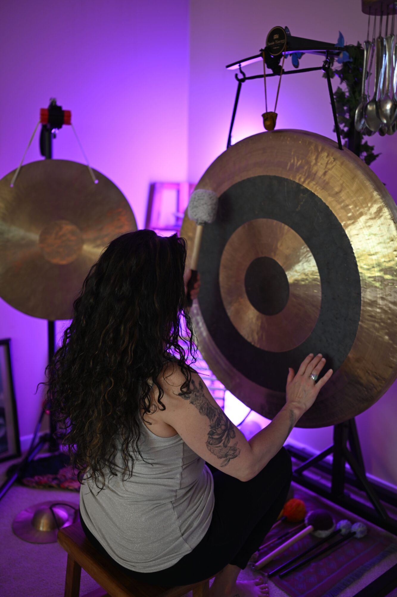 Person with long dark hair and tattoos playing a large cymbal in a music studio with purple lighting.