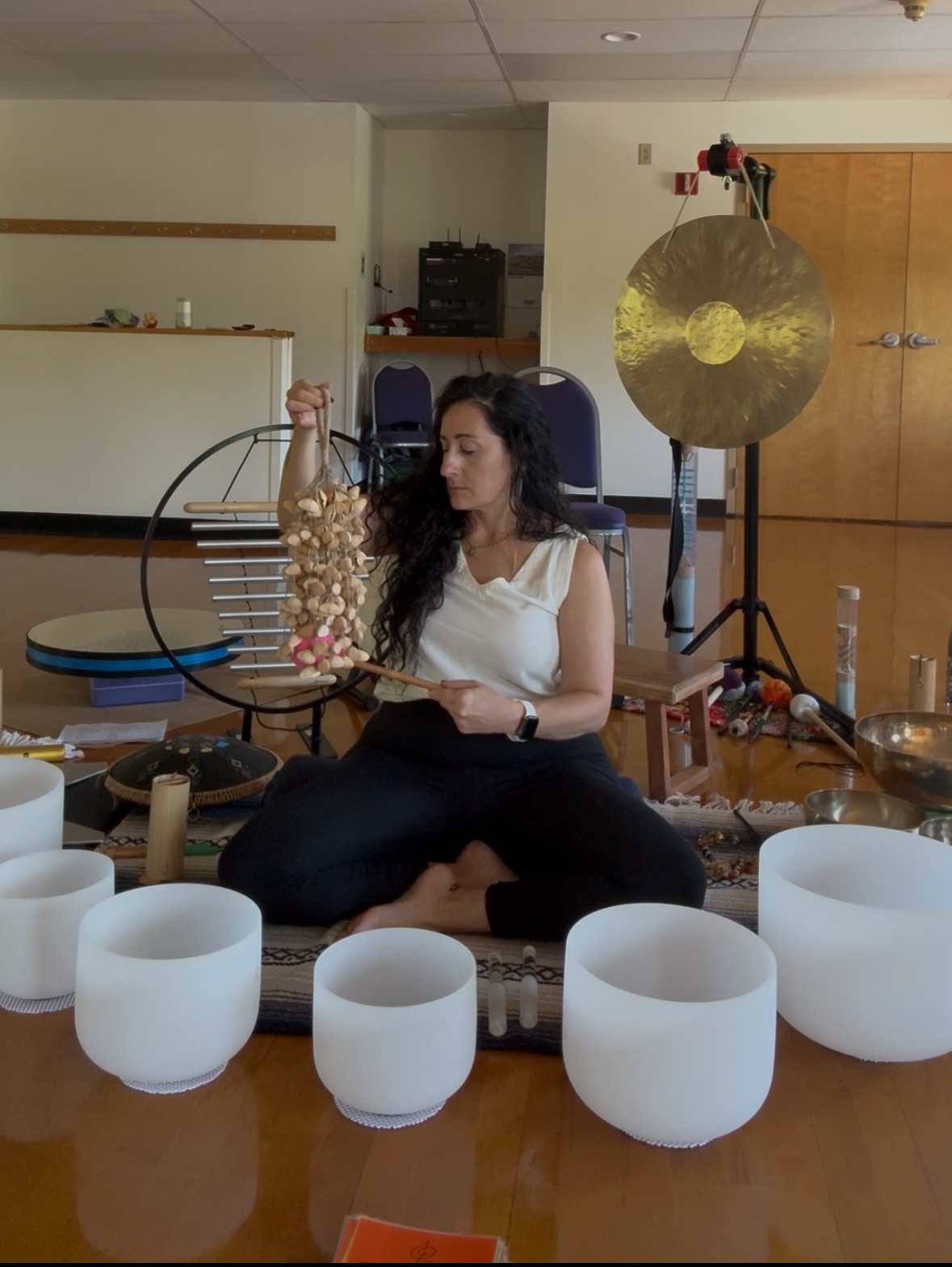 Woman sitting cross-legged on floor, playing crystal bowls, with a gong and other instruments in background.