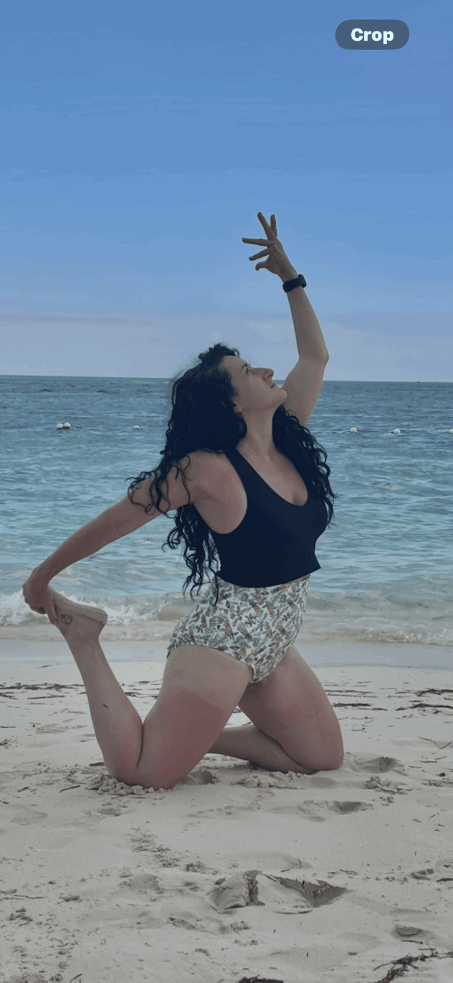 Person performing a dance pose on beach with ocean and sky background, one arm raised, knee on sand, wearing black top and patterned shorts.