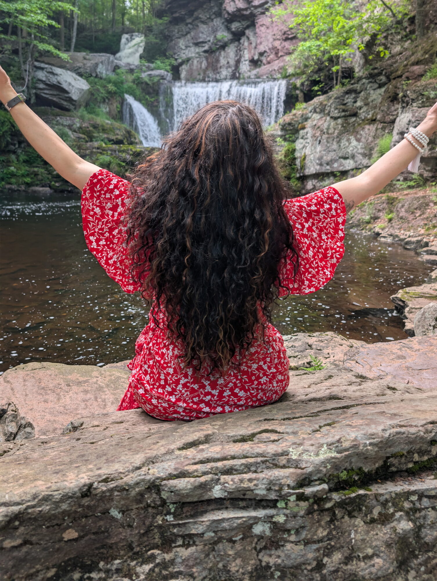 Person with long curly hair sitting on a rock near a waterfall with arms outstretched, wearing a red floral dress.