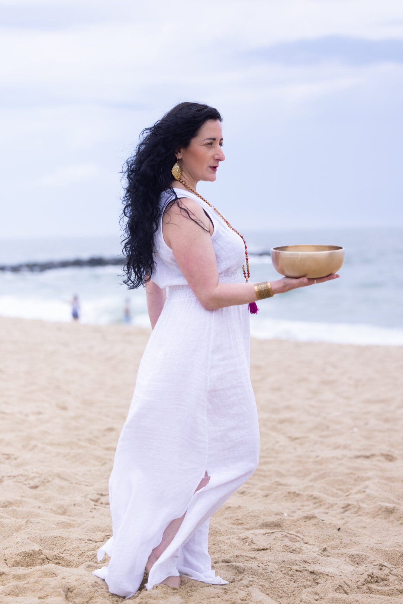 Woman in white dress holding a bowl on a sandy beach with ocean and sky in background.