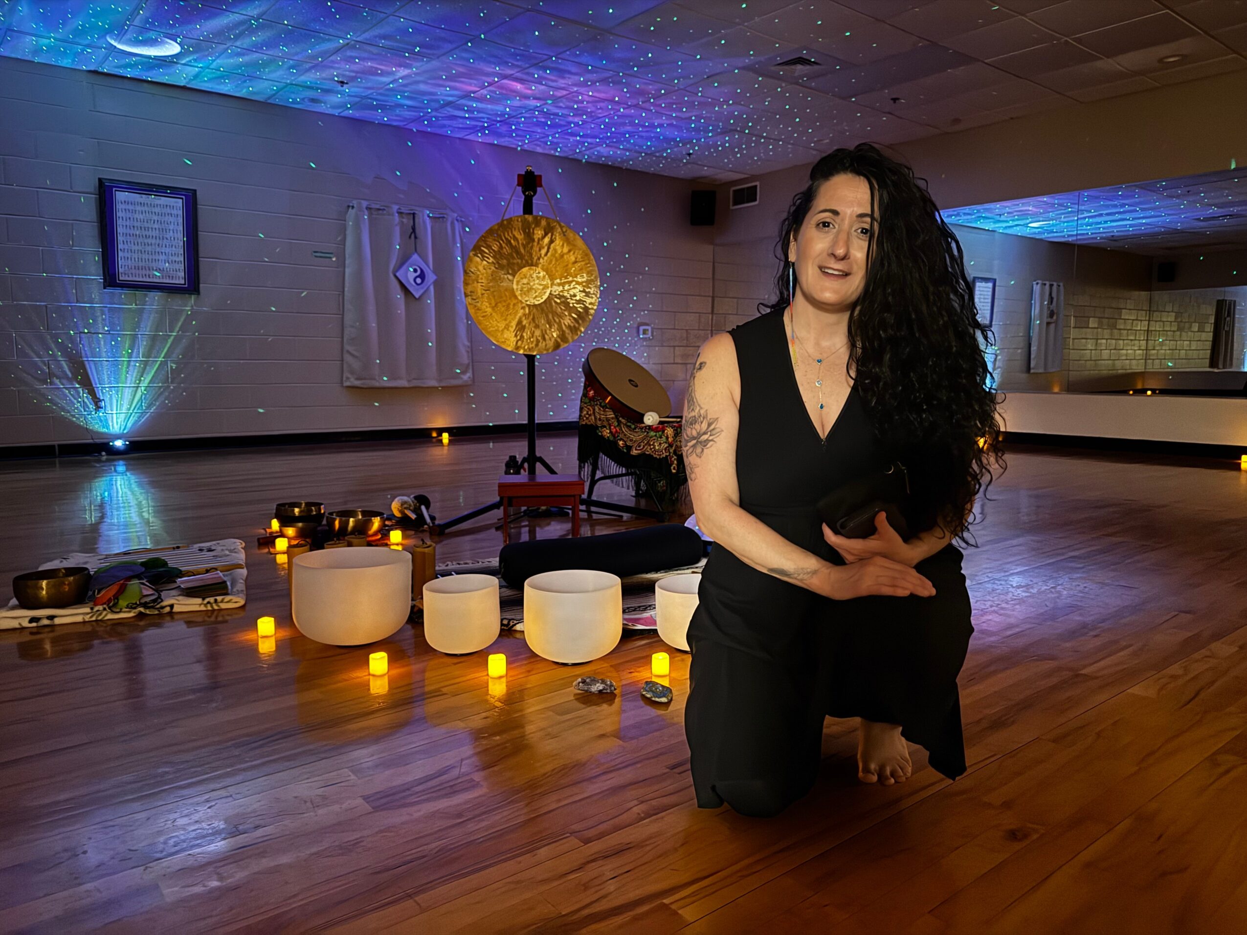 Woman with long curly hair kneeling on a wooden floor in a dimly lit room with candles and decorative elements.