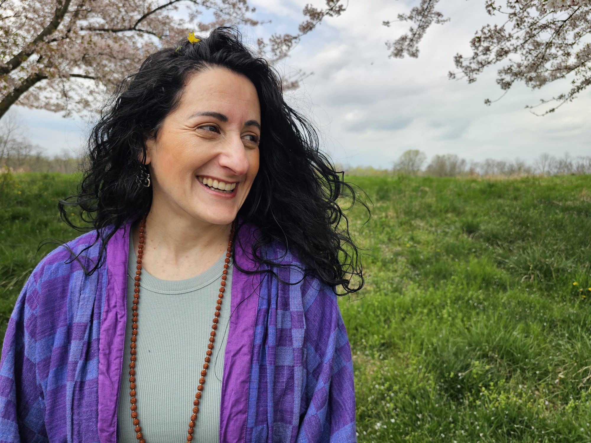 Woman with curly black hair smiling outdoors, wearing a purple jacket and light blue top, with trees and grass in background.