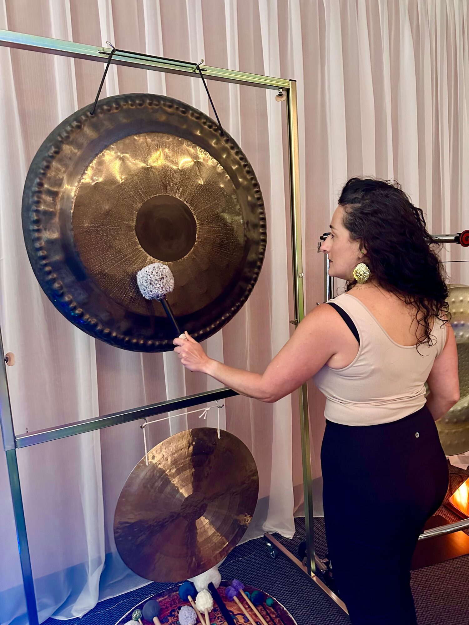 Woman strikes a gong with a mallet, standing in front of a large hanging gong and a smaller one below, with curtains in the background.