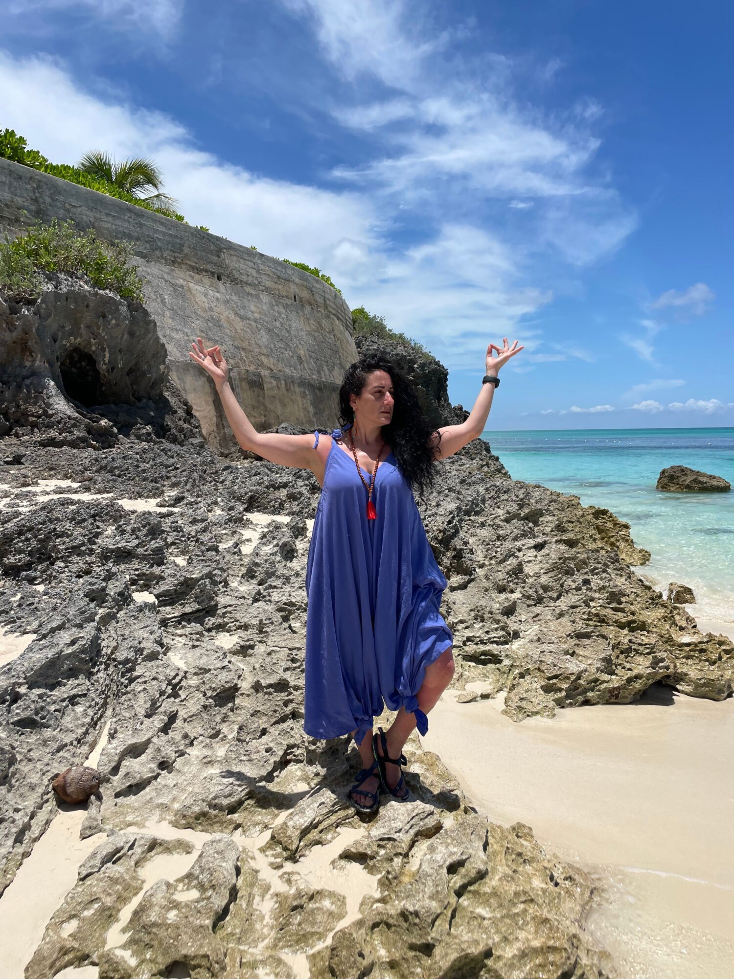 Person in a blue dress standing on rocks at the beach, with ocean and sky in the background.