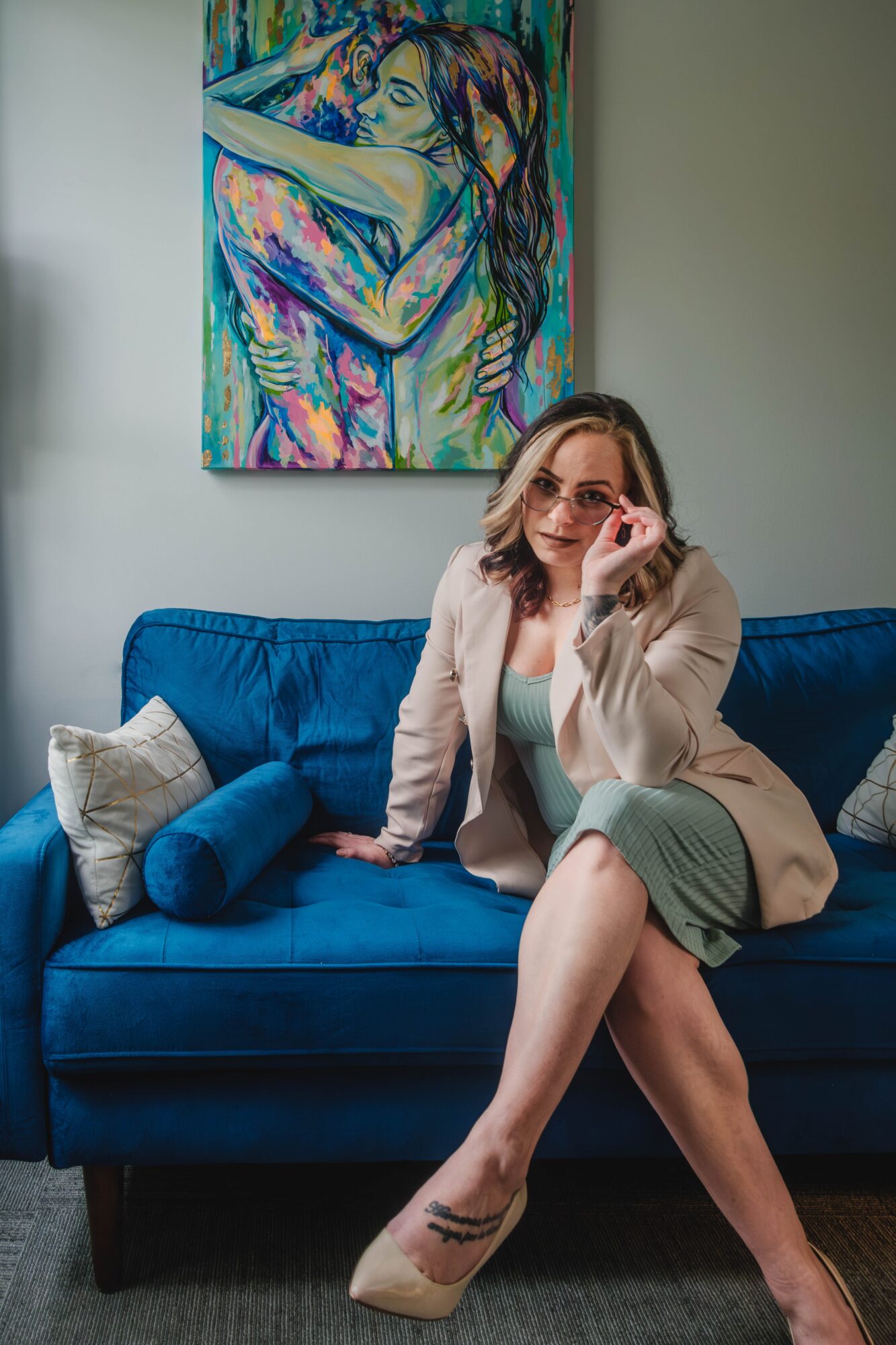 Woman with glasses sitting on a blue sofa, holding her glasses, with a colorful painting of two people hugging on the wall behind her.