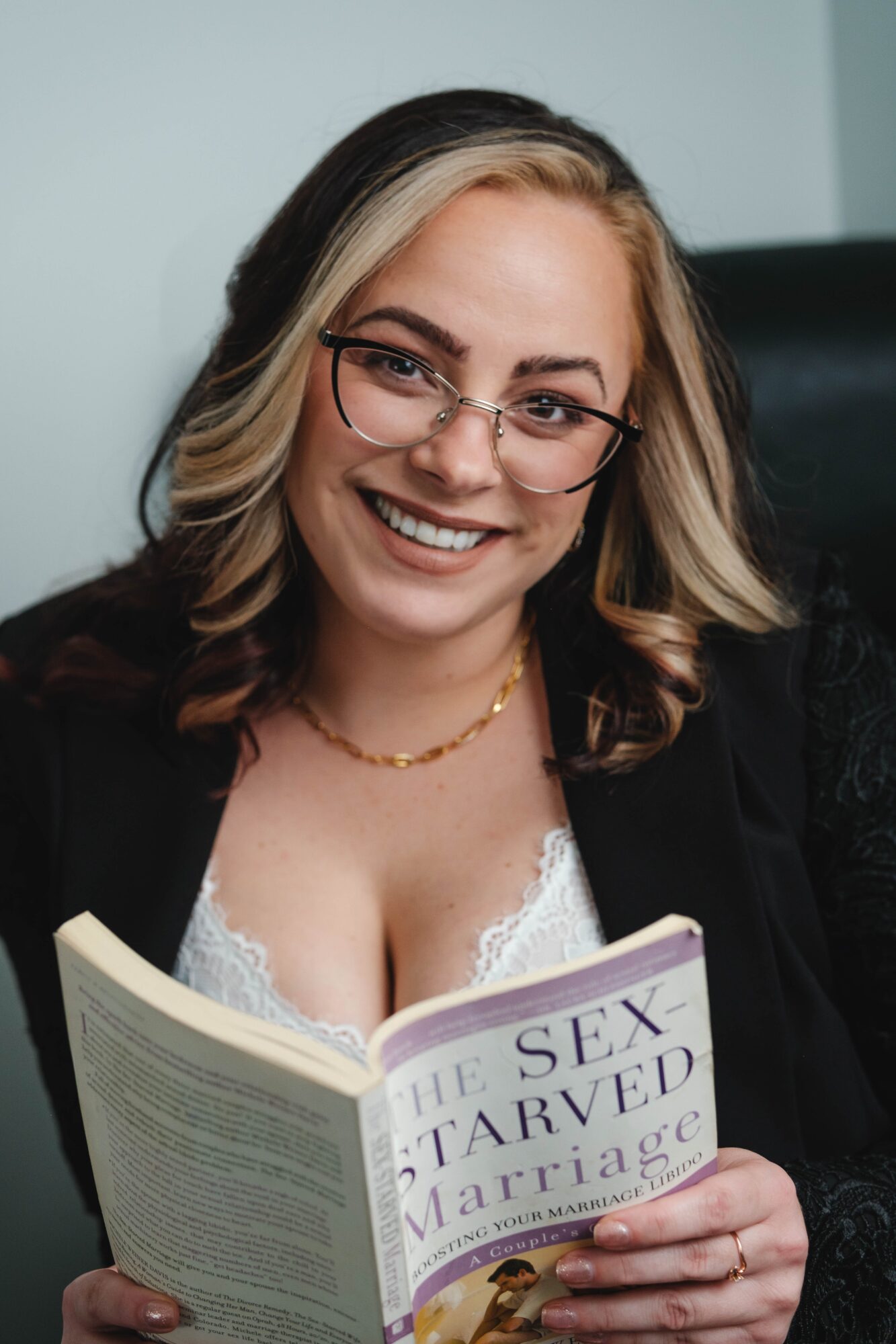 Woman with glasses smiling, holding a book titled 'The Sex Starved Marriage,' sitting in a chair.