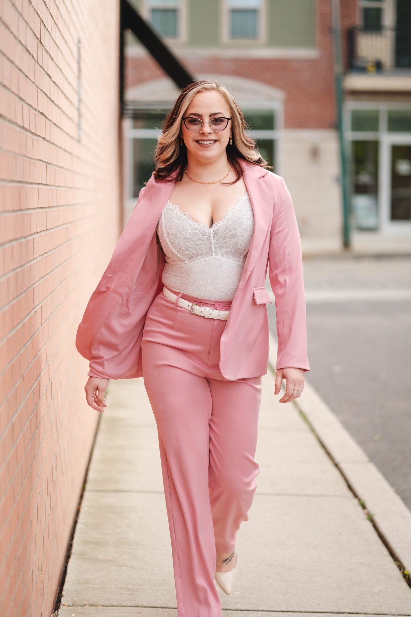 Young woman with glasses walking on sidewalk, wearing pink blazer and pants, smiling, in urban setting.