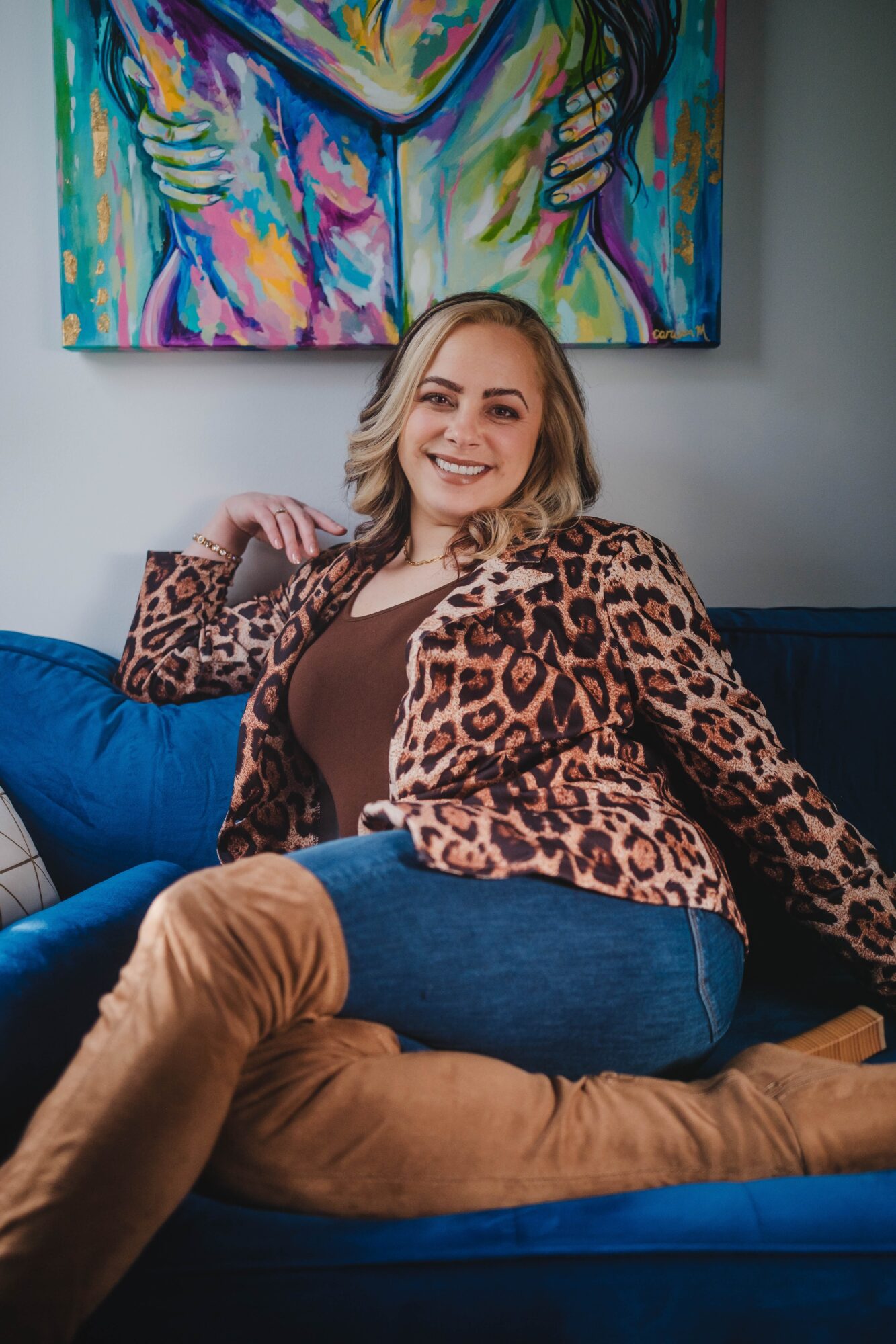 Woman with shoulder-length hair smiling, sitting on a blue couch, wearing a leopard print jacket and brown pants, in front of colorful artwork.