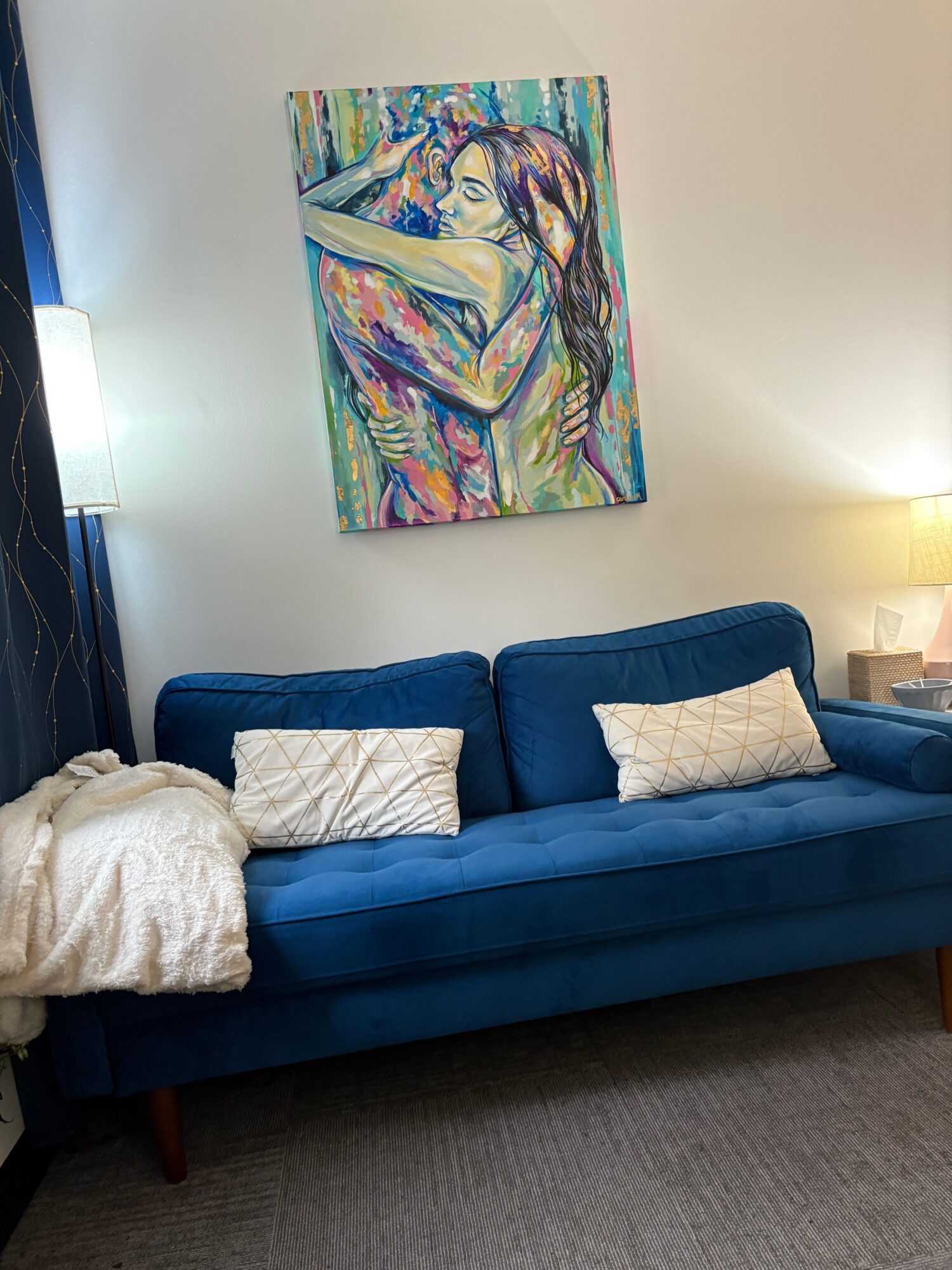 Blue sofa with three white pillows in a room with a colorful painting on the wall.