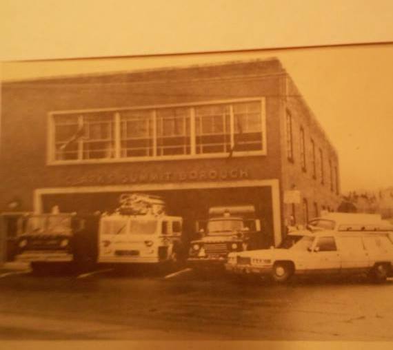 Vintage brick building with large windows and parked cars in front, sepia tone, from an earlier era.