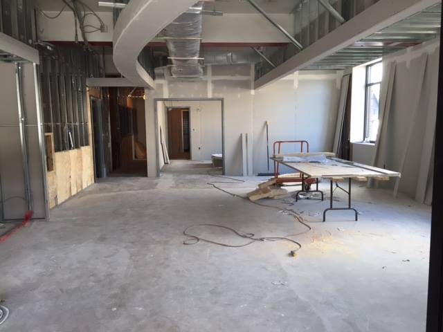 Construction site with unfinished floor, tables, and building materials, large window, and exposed ceiling ducts.