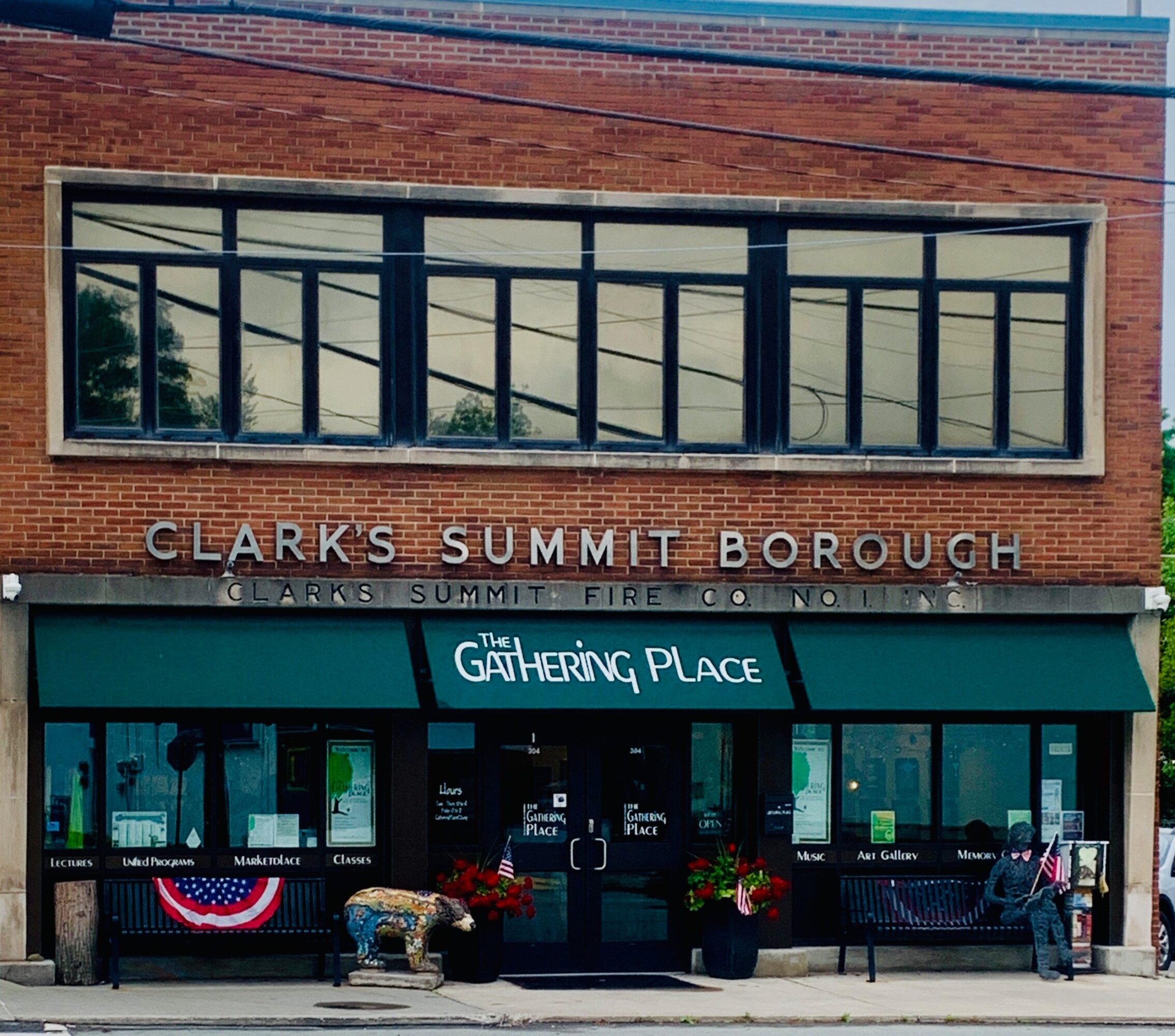 Brick building with large windows and a storefront with a green awning, flowers, and a dog statue outside.