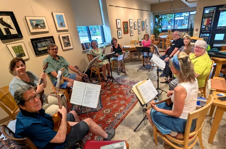 Group of people sitting in a circle with music stands, in a room decorated with framed pictures and a rug.