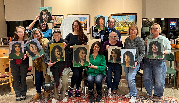 Group of people holding portrait paintings in an art gallery, smiling, with paintings displayed behind them.