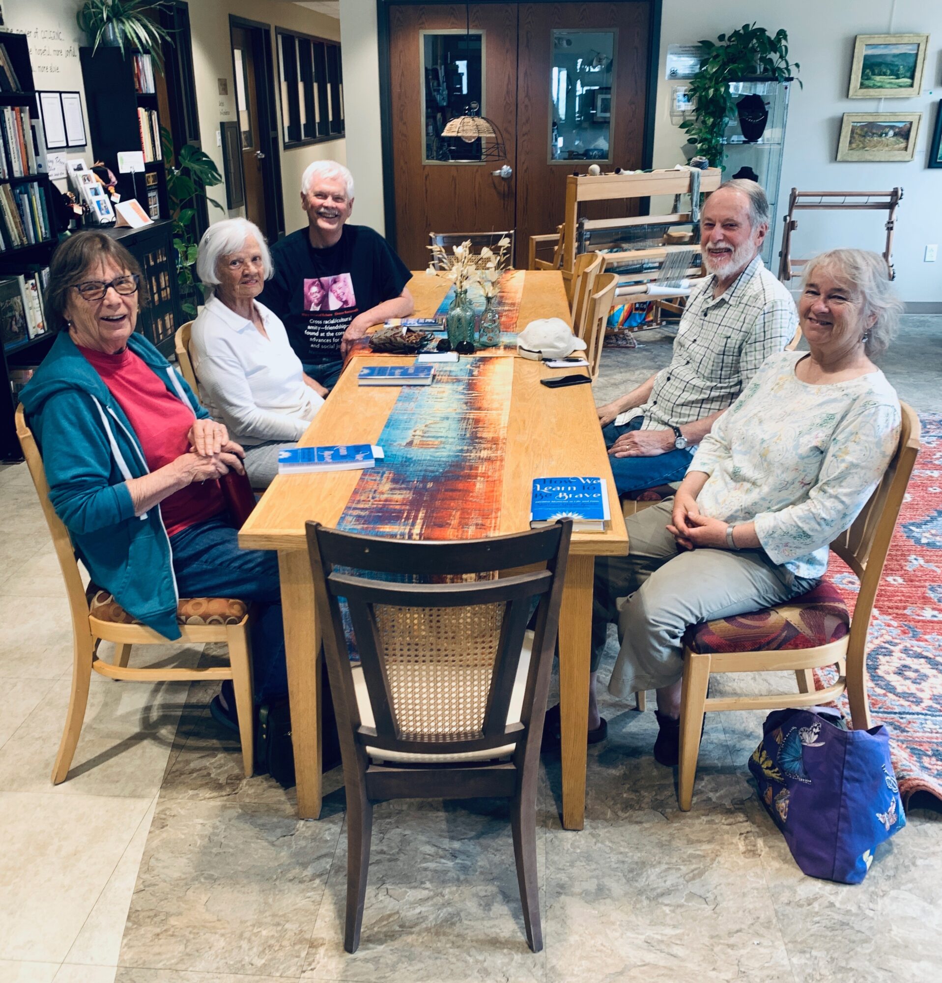 Five people sit around a wooden table in a room with bookshelves, artwork, and a rug, smiling at the camera.