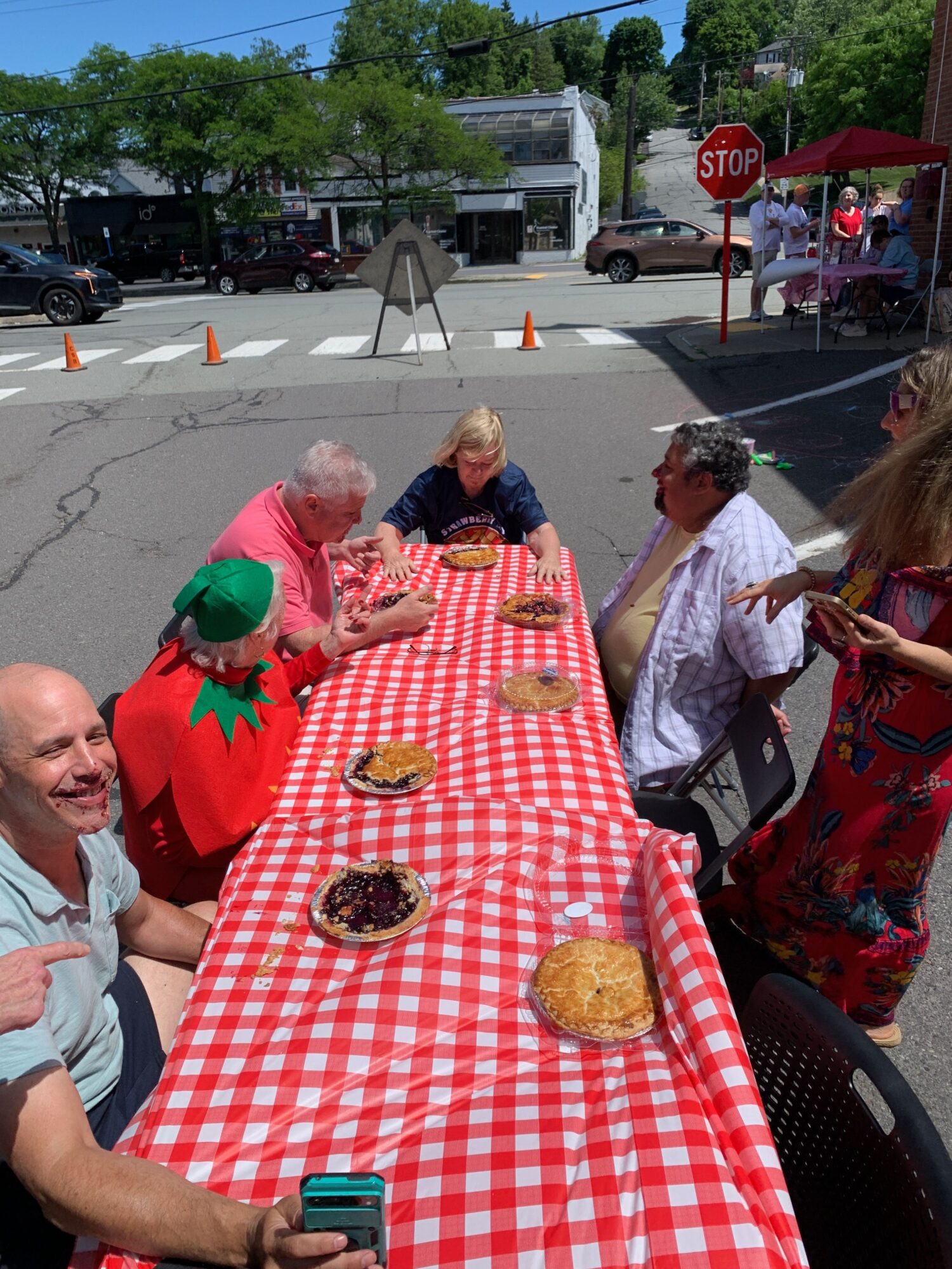 People gather around a table with a red and white checkered tablecloth, enjoying food outdoors on a sunny day.