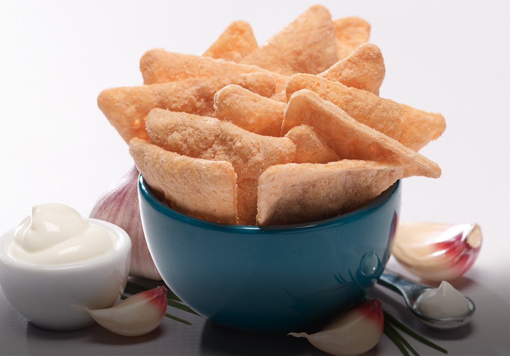 A blue bowl filled with fried chips, with garlic cloves and a small bowl of sauce nearby.