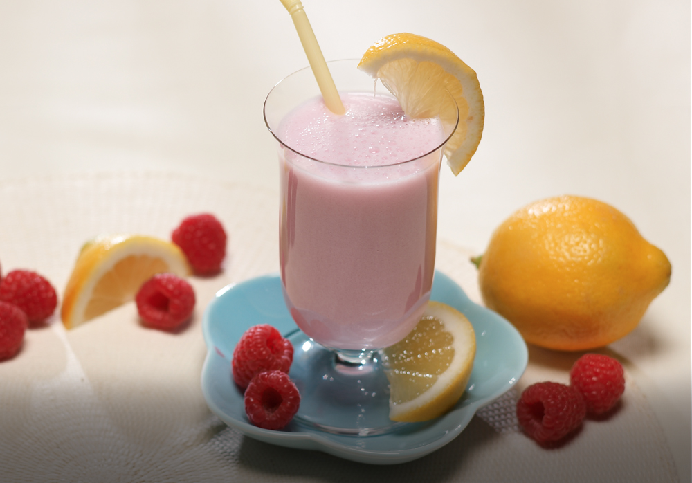 A pink beverage in a glass with lemon slice and straw, surrounded by raspberries, lemon, and lemon slices on a blue plate.