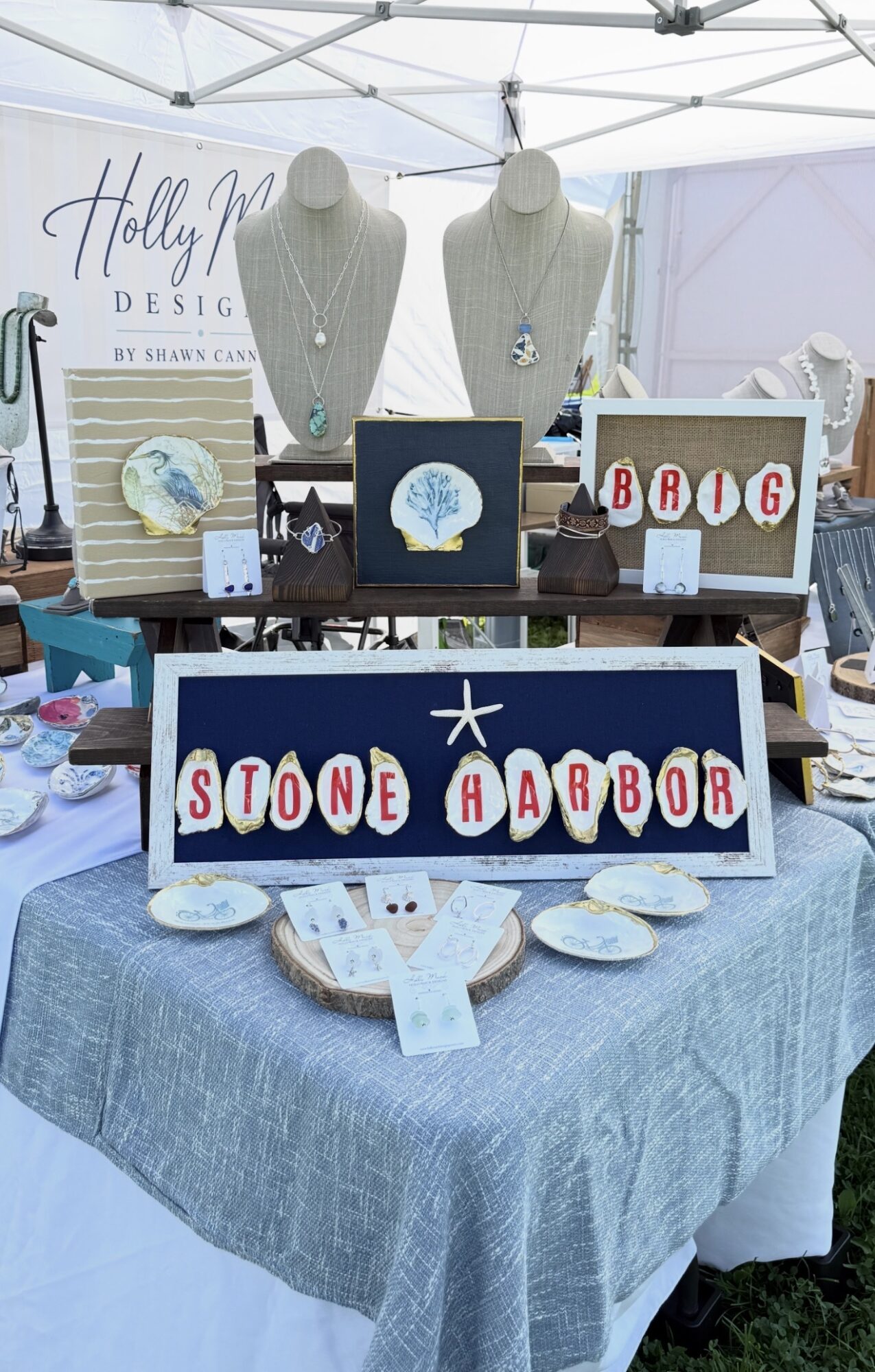 Display table with jewelry, signs, and decorative items at an outdoor event, under a canopy.