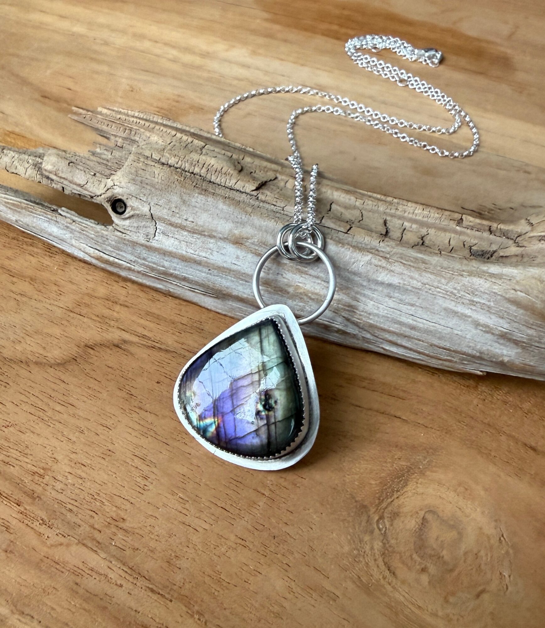 Silver necklace with a pendant featuring a landscape image, resting on a piece of driftwood on a wooden surface.