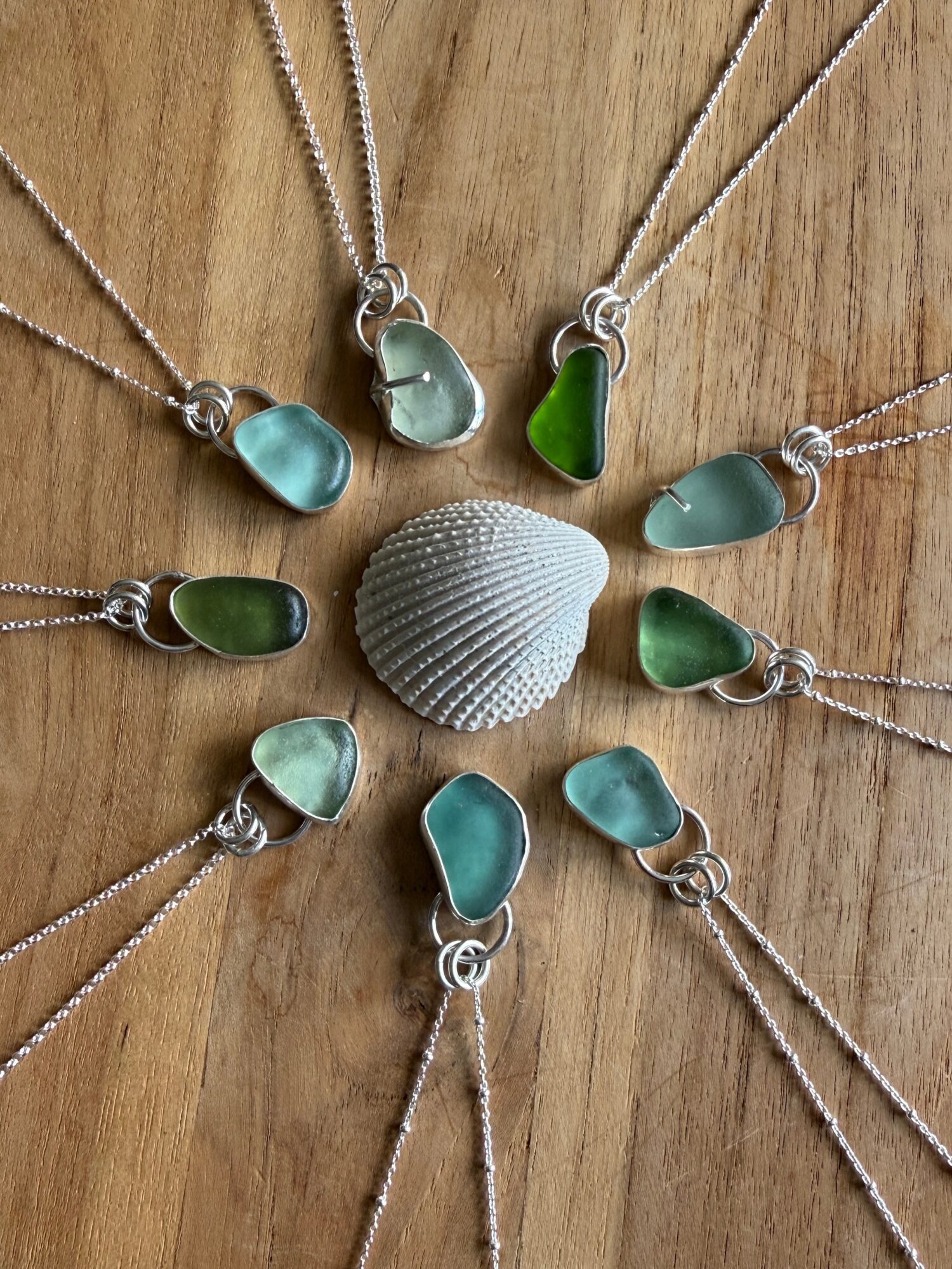 Collection of sea glass necklaces arranged around a seashell on a wooden surface, with various shades of green and blue glass.