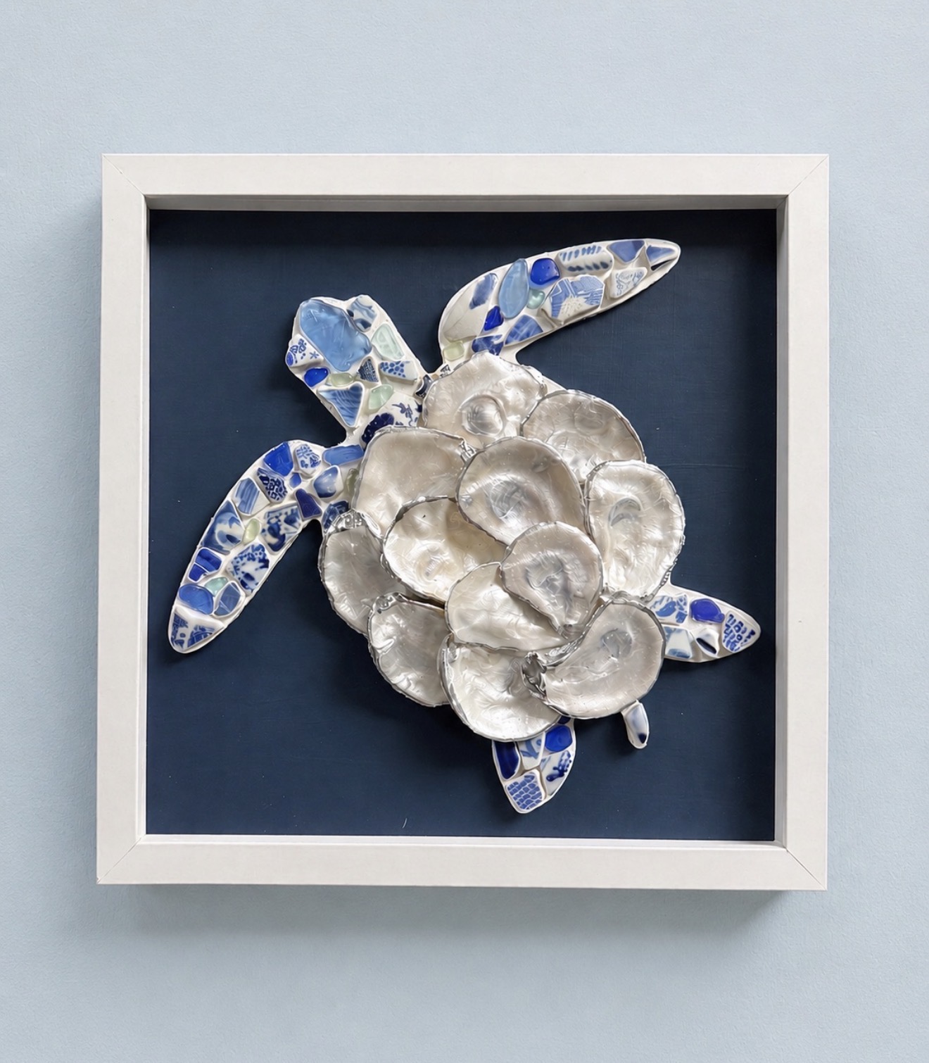 Framed artwork of a turtle with a shell made of shiny, metallic shells and limbs with blue mosaic patterns, on a blue background.