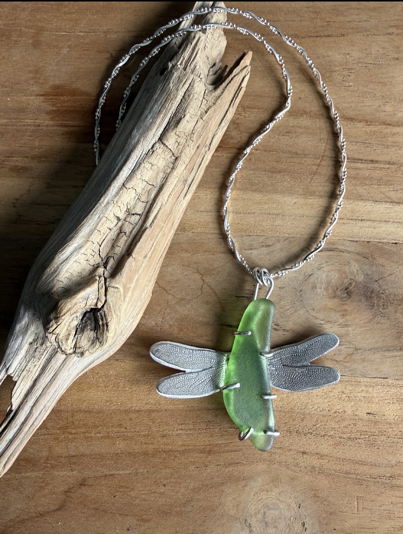Necklace with a green dragonfly pendant on a wooden surface, next to a piece of driftwood.