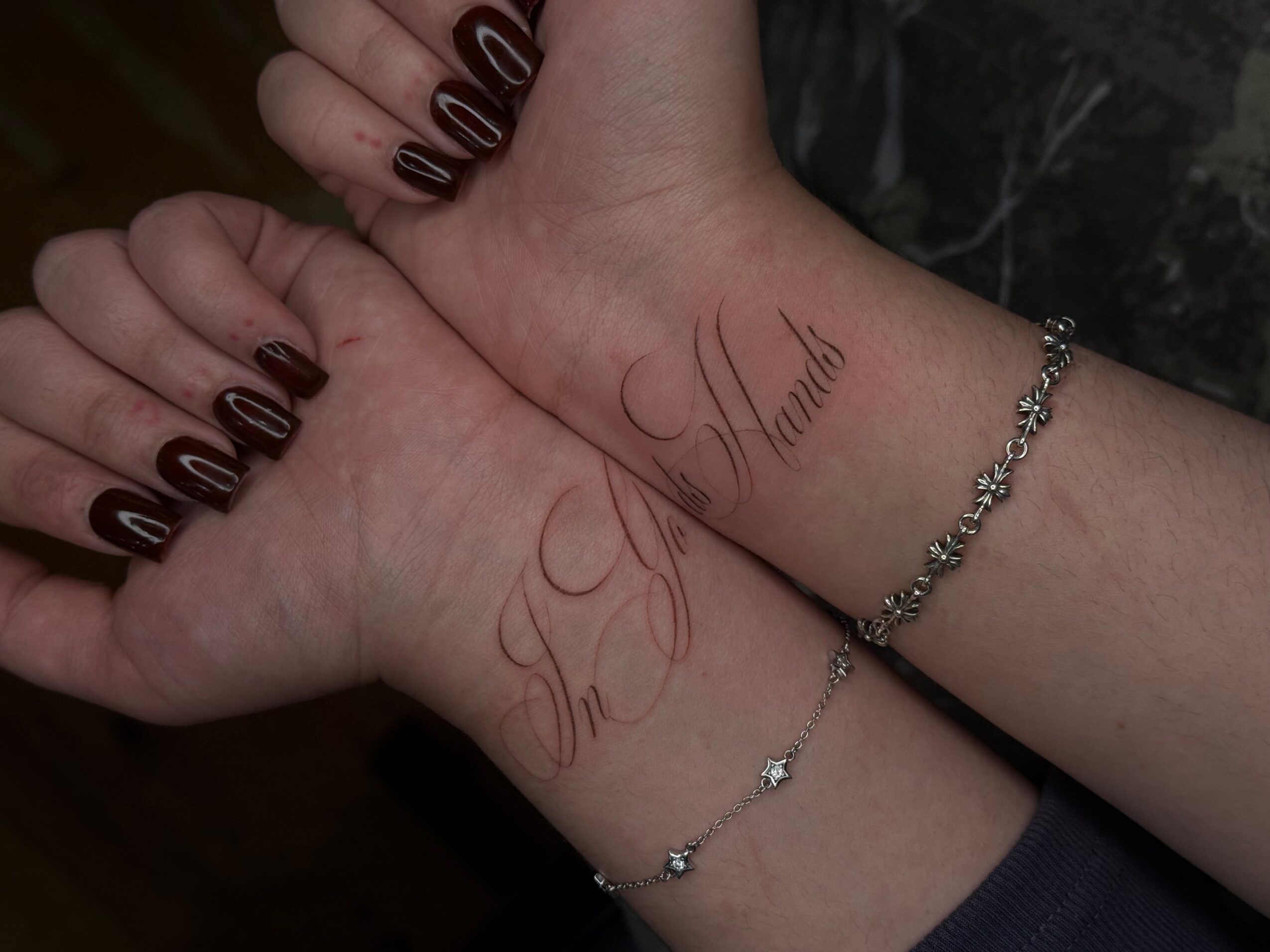 Two wrists with tattoos and jewelry, one with black nail polish, in a close-up shot.