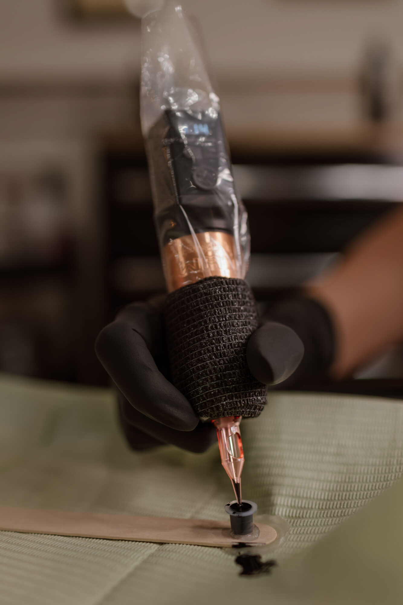 Person wearing black glove holding a tattoo machine, tattooing on skin, background blurred.
