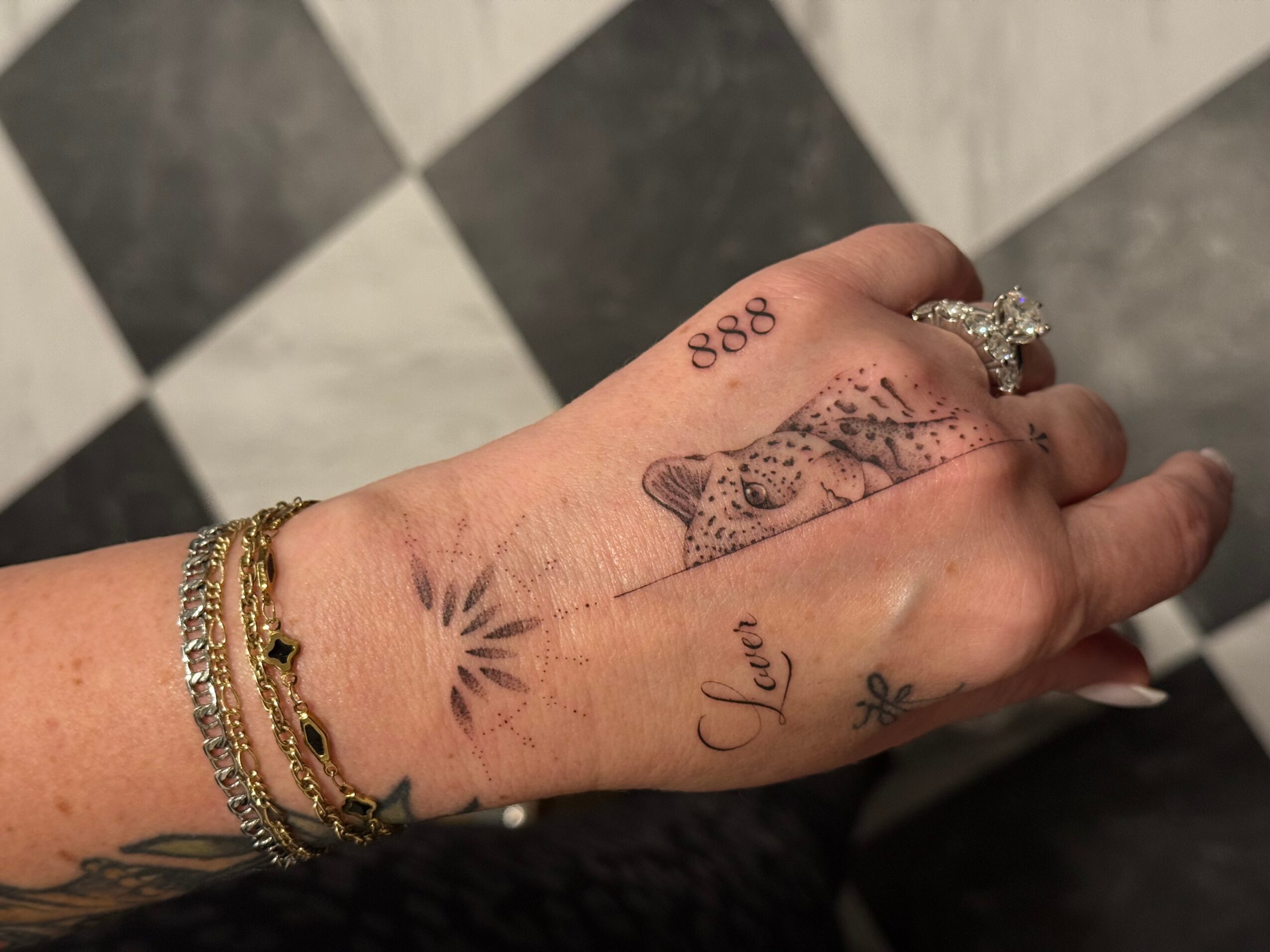 Hand with multiple tattoos, rings, and bracelets, showing a fish, the word 'Love', and the number '888'.