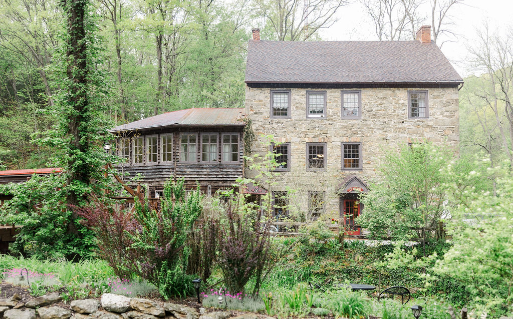 Stone house with a wooden porch, surrounded by trees and greenery, in a lush garden setting.