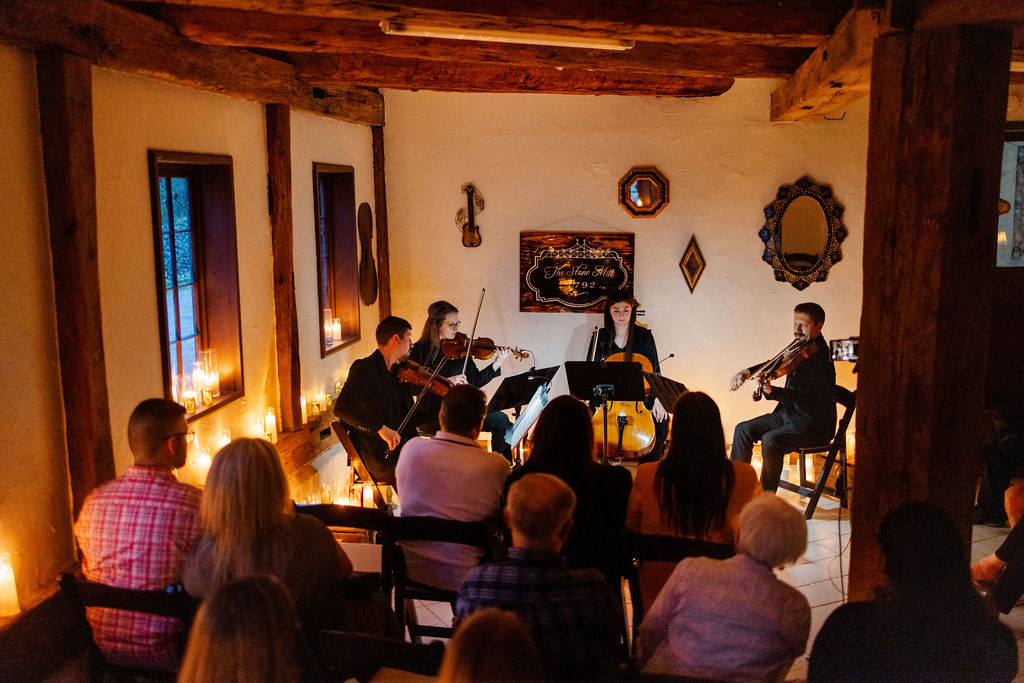 Group of musicians performing on a small stage with an audience seated in a cozy room, warm lighting, wooden beams, and decorative wall items.