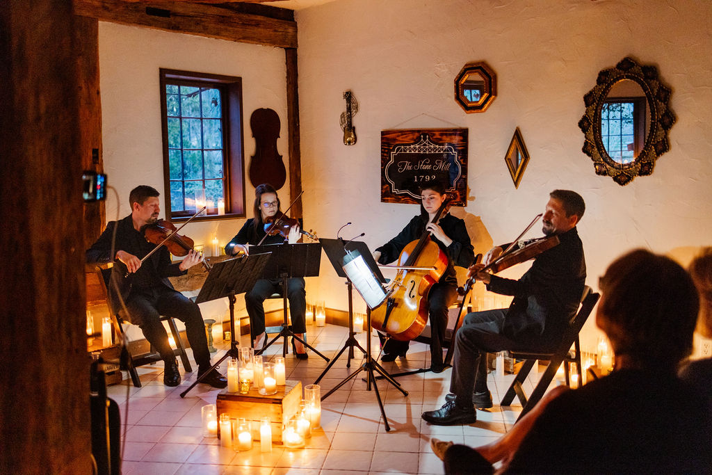 Four musicians perform in a cozy room with candles, a window, and decorative mirrors on the wall.