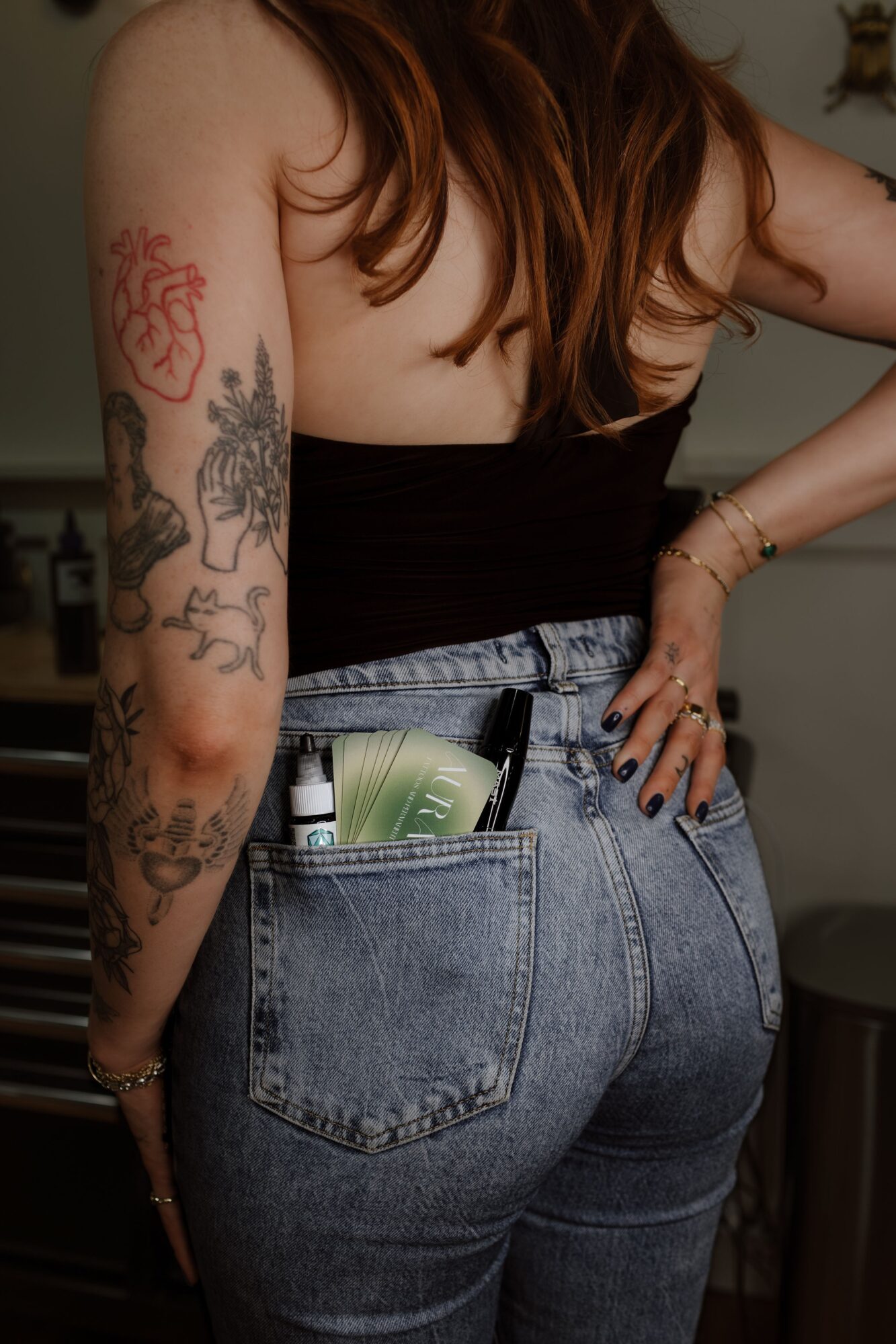 Person with tattoos on arm, wearing a black top and high-waisted jeans, with a hand on hip, in a room.
