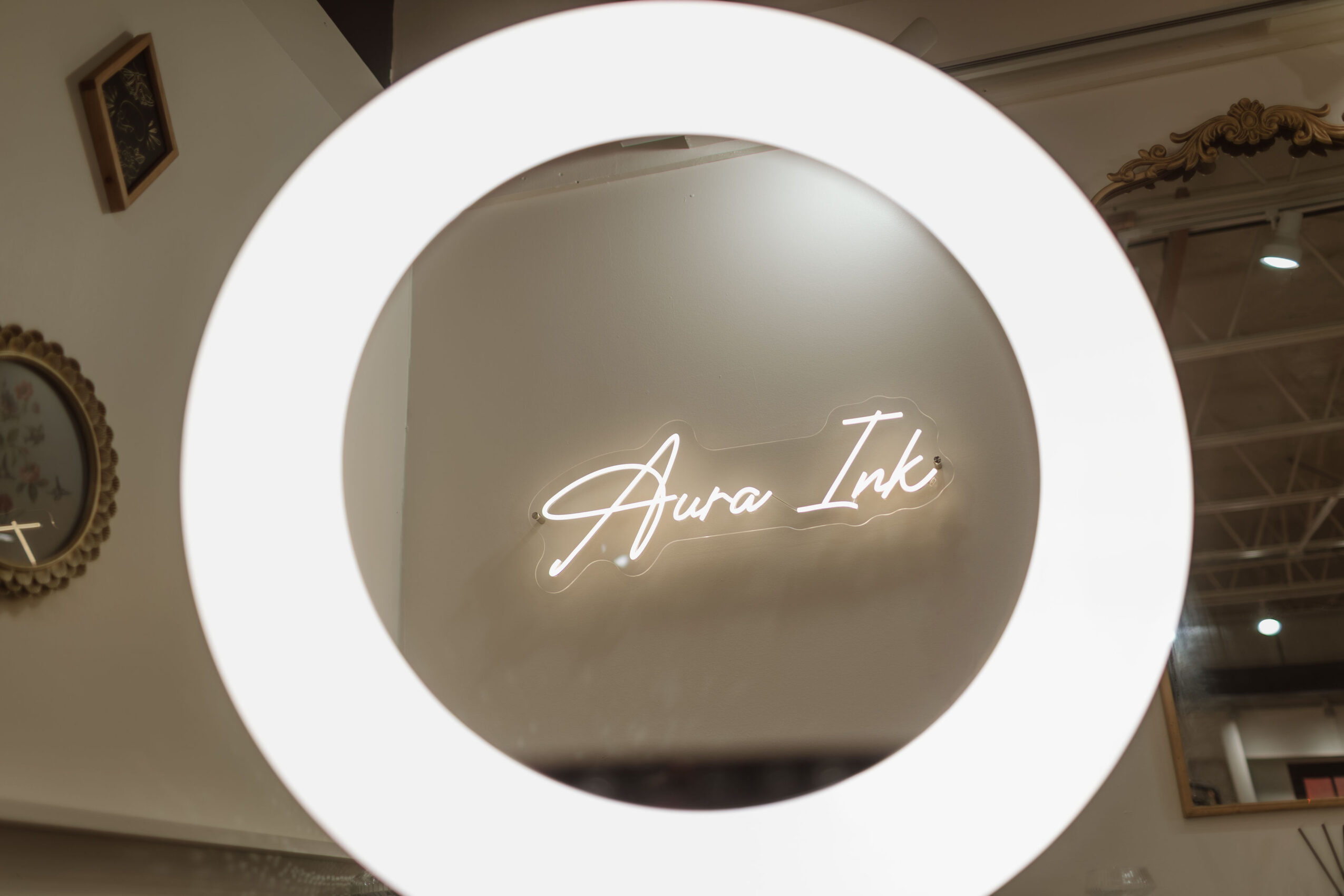 Neon sign reading 'Aura Ink' on a wall, viewed through a circular mirror or frame, with part of a clock and framed picture visible.
