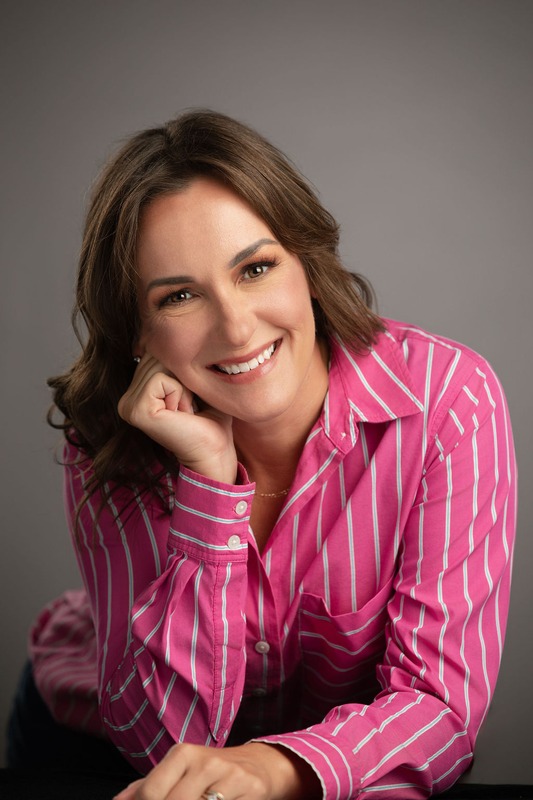 Woman with shoulder-length brown hair smiling, resting her chin on her hand, wearing a pink and white striped shirt, gray background.