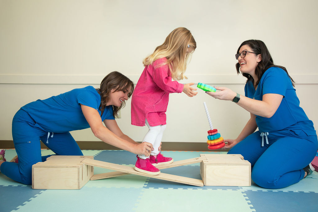 Two women in scrubs and a young girl are playing on a balance beam with toys, smiling and engaging with each other.