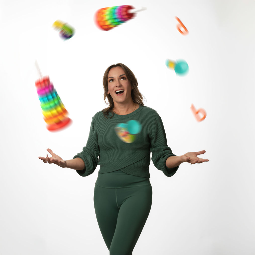 Woman in green outfit juggling colorful objects, smiling, against a white background.