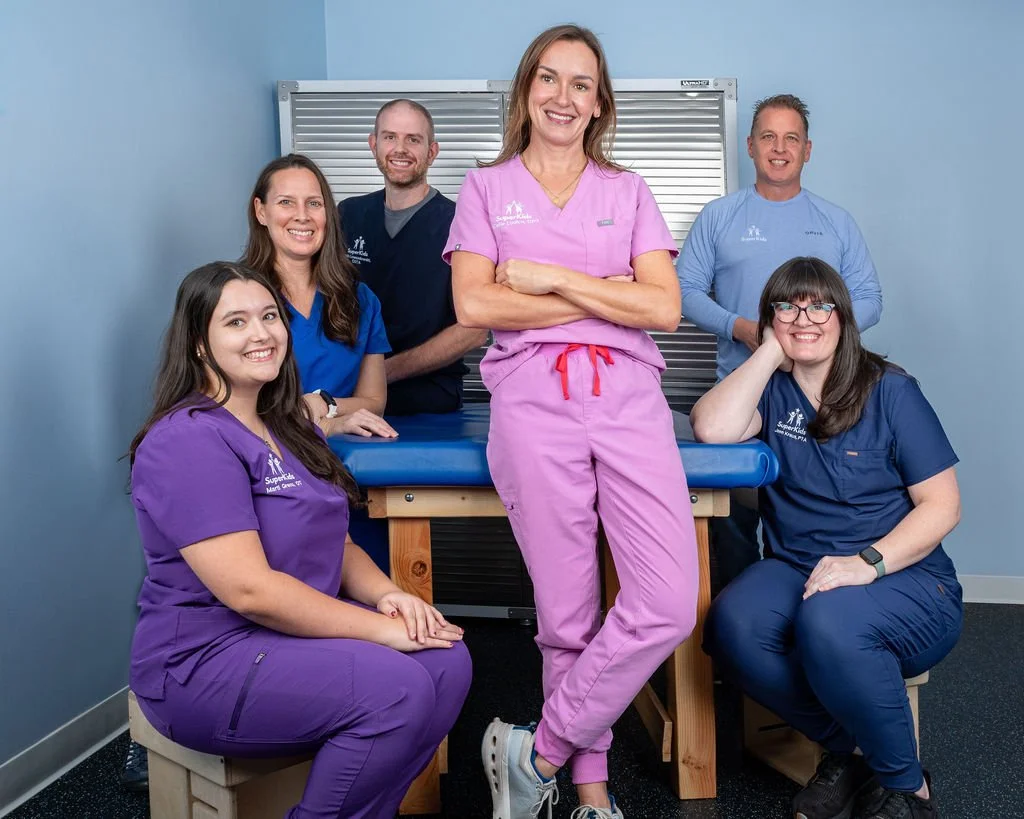 Group of six healthcare professionals, five women and one man, in scrubs and lab coat, smiling in a clinical setting.