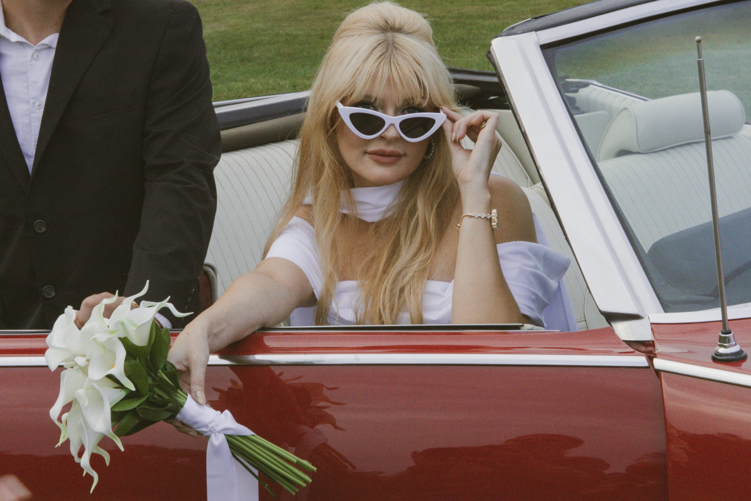 Woman with long blonde hair and white sunglasses sitting in a red convertible car, holding a bouquet of white flowers.