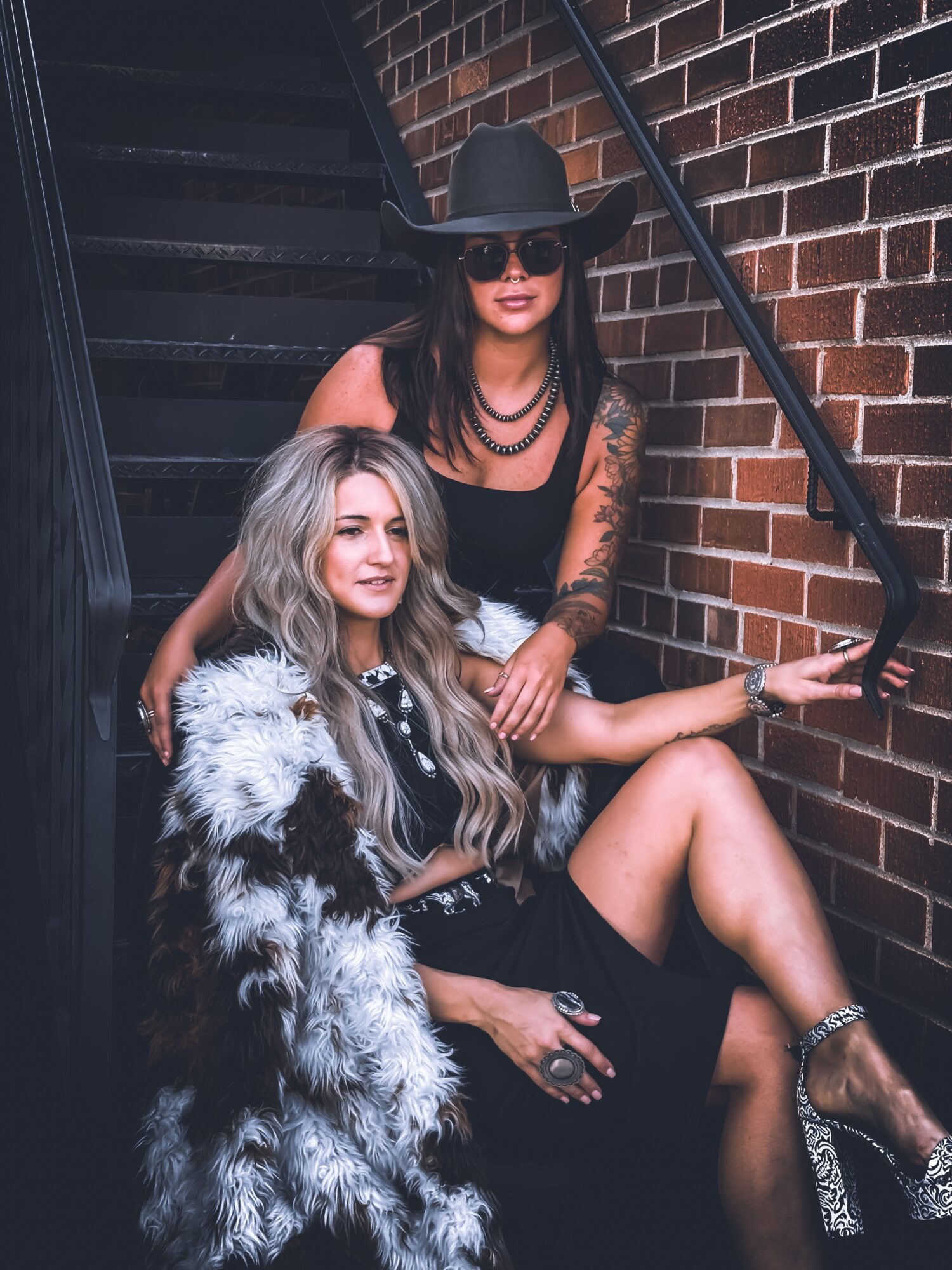Two women sitting on stairs against a brick wall, one wearing a large hat and sunglasses, the other with long wavy hair and a fur coat.