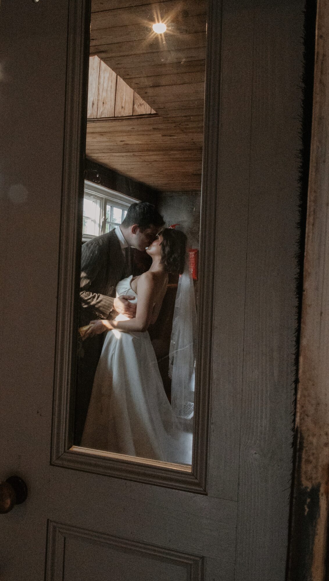Two people, a man and a woman, are taking a selfie in a mirror, with a wooden ceiling visible above them.
